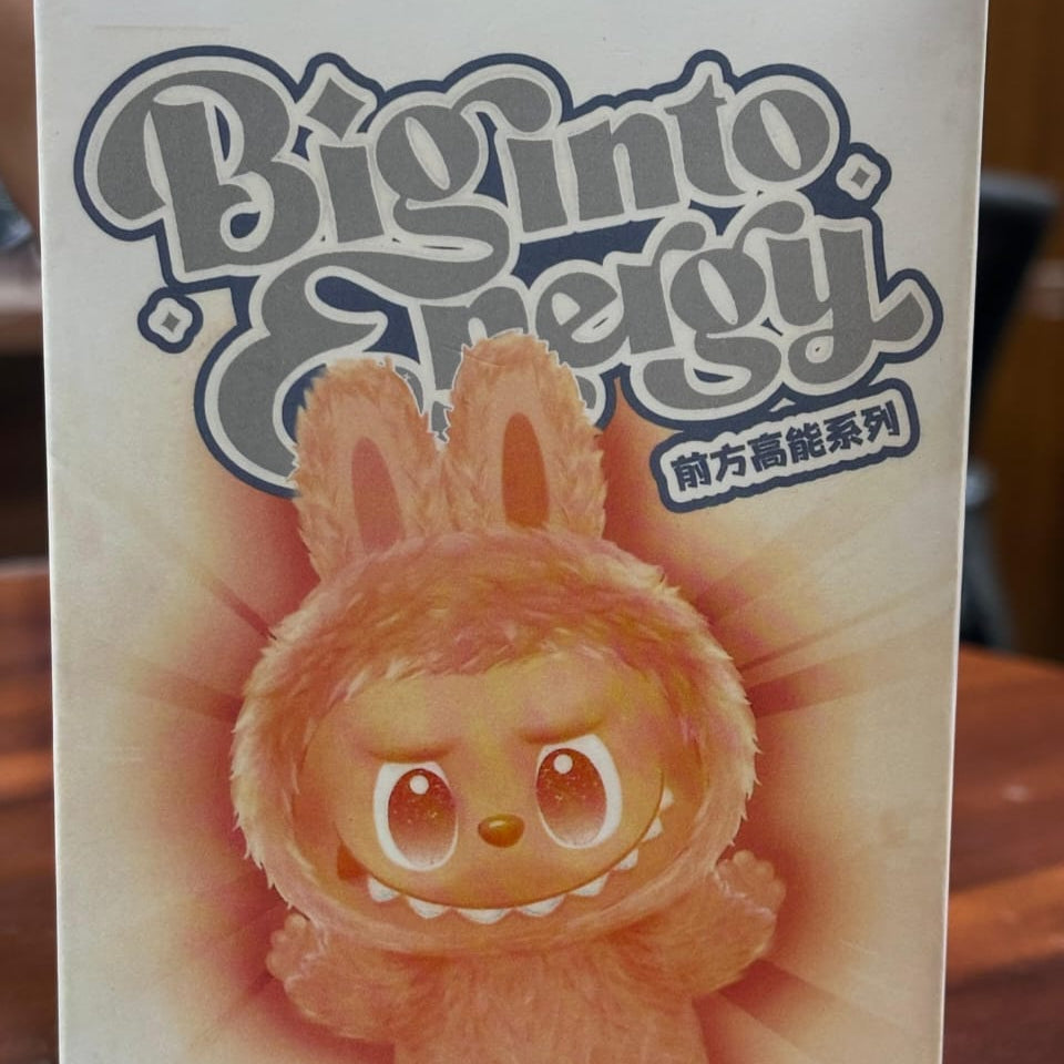 Box with a plush toy resembling a character from 'Big Into Energy' on a wooden surface.